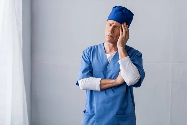 Tired and middle aged doctor in blue scrub hat touching head on grey — Stock Photo