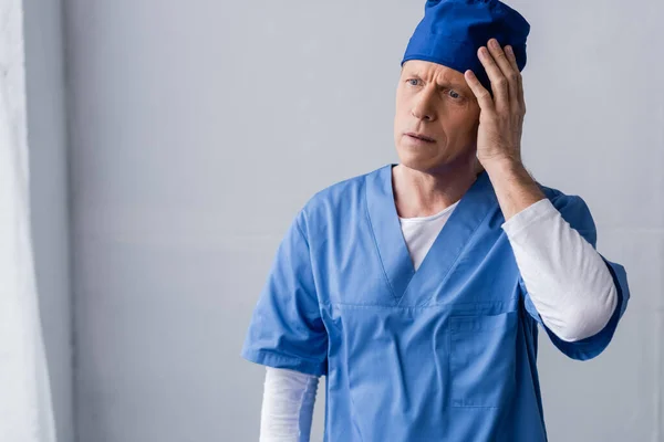 Exhausted and middle aged doctor in blue scrub hat touching head on grey — Stock Photo
