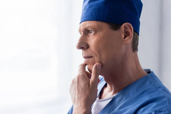 Médico de meia-idade pensativo em esfregar chapéu tocando rosto enquanto pensa no branco — Fotografia de Stock