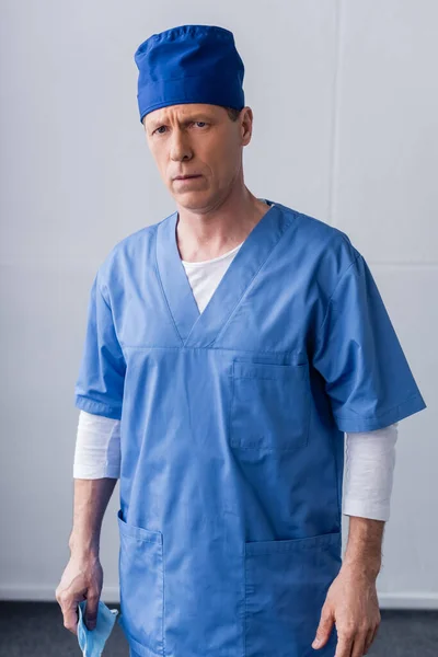 Maturo medico in blu scrub cappello che tiene maschera medica su grigio — Foto stock