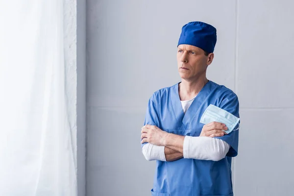 Задумчивый и зрелый доктор в голубой шапке, стоящий со скрещенными руками и держащий медицинскую маску на сером — стоковое фото