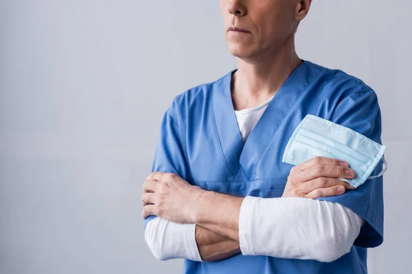 Cropped view of middle aged doctor standing with crossed arms and holding medical mask on grey — Stock Photo