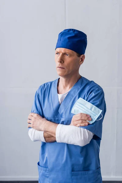 Middle aged doctor in blue scrub hat standing with crossed arms and holding medical mask on grey — Stock Photo