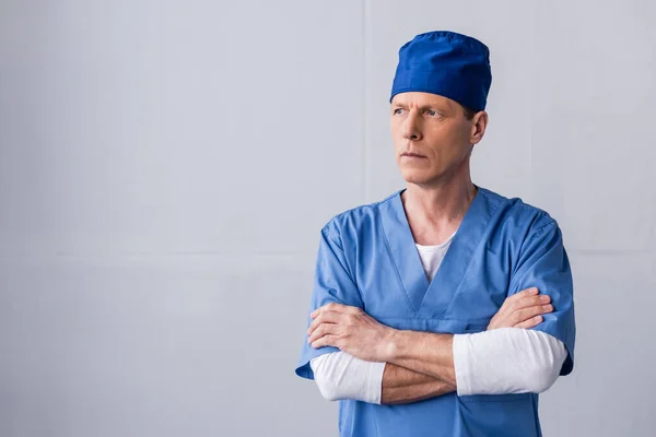Pensivo médico de meia-idade em chapéu de esfoliação azul de pé com braços cruzados em cinza — Fotografia de Stock