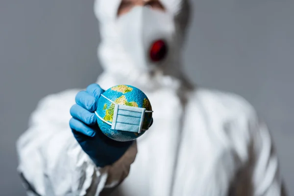 Selective focus of man in hazmat suit holding small globe in medical mask isolated on grey — Stock Photo