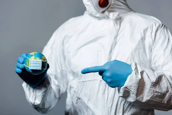 Cropped view of man in hazmat suit pointing with finger at small globe in medical mask isolated on grey — Stock Photo