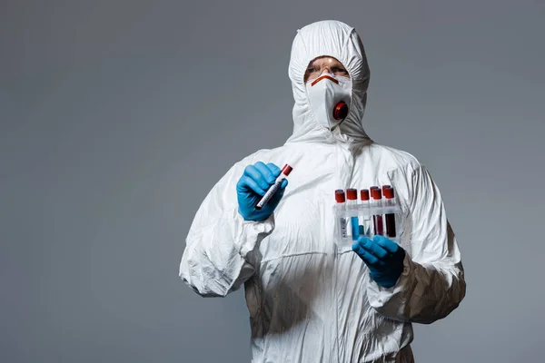 Mature doctor in hazmat suit, medical mask and goggles holding test tube with coronavirus lettering isolated on grey — Stock Photo