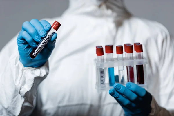 Cropped view of mature doctor in hazmat suit and latex gloves holding test tube with coronavirus lettering isolated on grey — Stock Photo