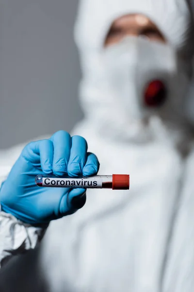 Foyer sélectif du médecin mature en combinaison hazmat, masque médical et lunettes tenant le tube à essai avec lettrage coronavirus isolé sur gris — Photo de stock