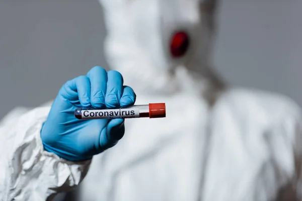 Vista recortada del hombre en traje de materiales peligrosos y guante de látex que sostiene el tubo de ensayo con letras de coronavirus aisladas en gris - foto de stock