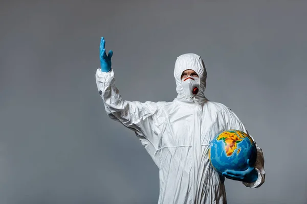 Зрілий лікар у костюмі з туманом, медична маска та латексні рукавички, що тримають глобус та жестикулюють ізольовані на сірому — стокове фото