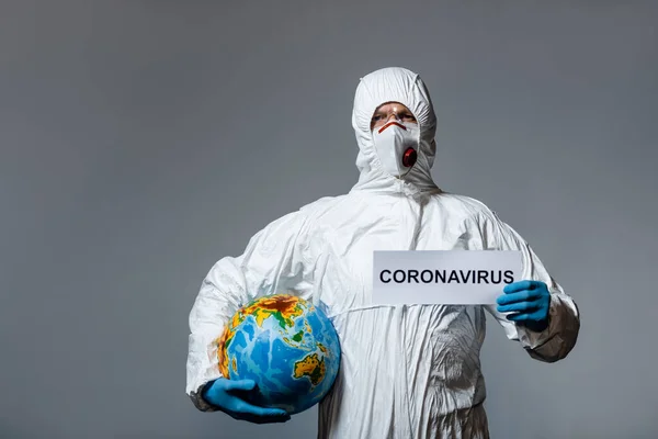 Mann in persönlicher Schutzausrüstung hält Globus und Papier mit Coronavirus-Schriftzug isoliert auf grau — Stockfoto