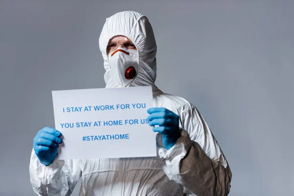 Medico maturo in tuta hazmat tenendo cartello con io rimango al lavoro per voi, si rimane a casa per noi lettering isolato su grigio — Foto stock