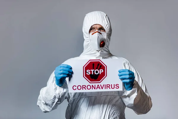 Зрілий лікар у костюмі з туманом, що тримає плакат із стоп-коронавірусом, записуючи ізольовані на сірому — стокове фото