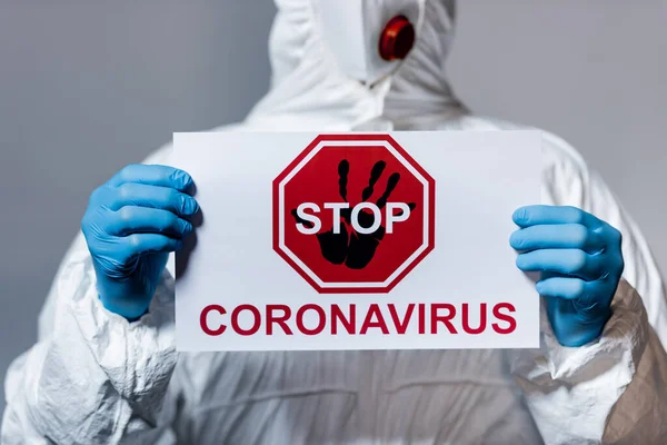 Vue recadrée du médecin adulte en combinaison Hazmat tenant une pancarte avec lettrage stop coronavirus isolé sur gris — Photo de stock