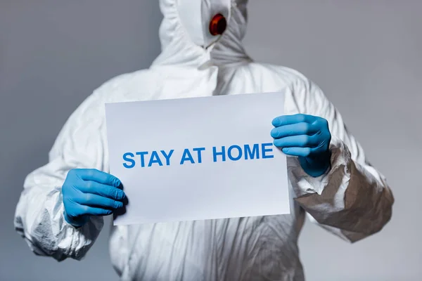 Vista cortada do homem em terno hazmat segurando cartaz com estadia em casa lettering isolado em cinza — Fotografia de Stock