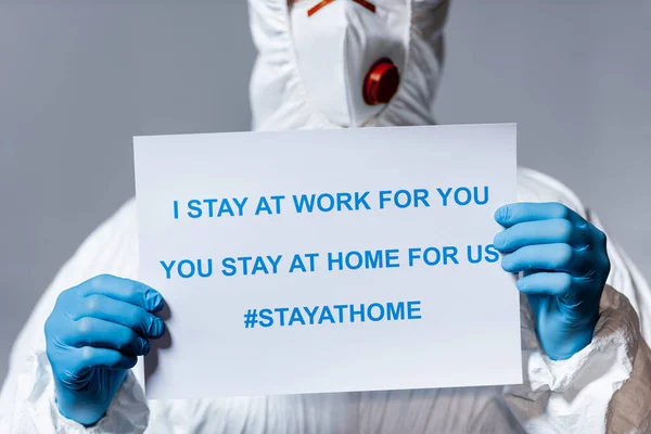 Cropped view of mature doctor in hazmat suit holding placard with i stay at work for you, you stay at home for us lettering isolated on grey — Stock Photo