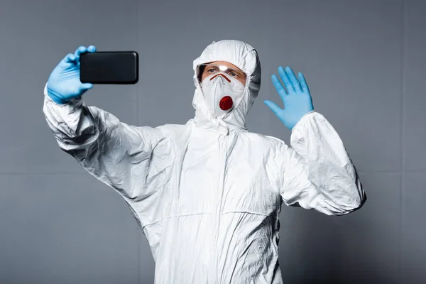 Man in personal protective equipment taking selfie and waving hand on grey — Stock Photo