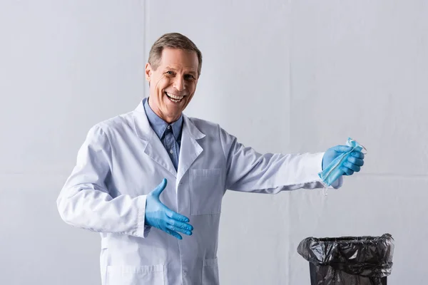 Happy mature doctor holding medical mask near trash can on white — Stock Photo