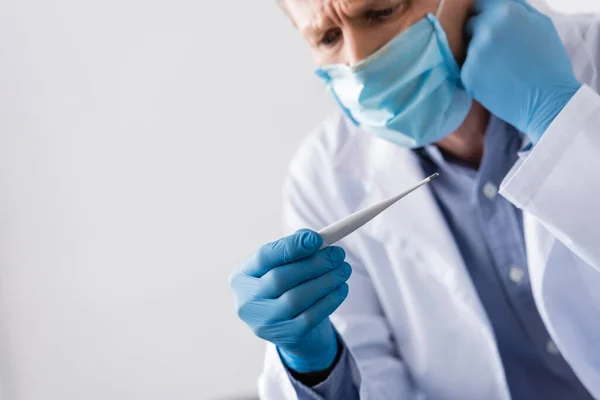 Selective focus of mature doctor in latex gloves looking at digital thermometer isolated on grey — Stock Photo