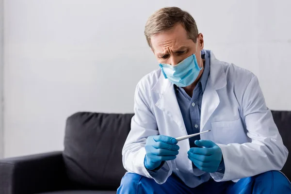 Mature doctor in medical mask and latex gloves looking at digital thermometer while sitting on grey — Stock Photo