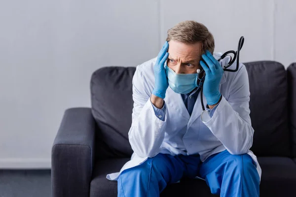 Müder, reifer Arzt mit Kopfschmerzen, der Stethoskop hält, während er auf dem Sofa sitzt — Stockfoto