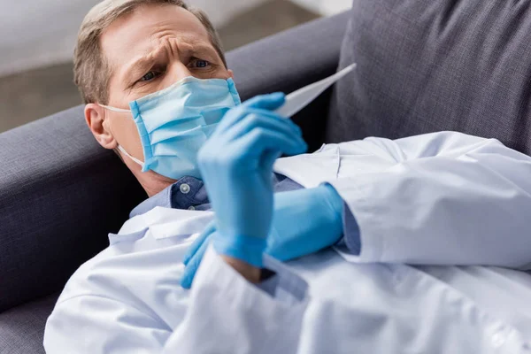Селективный фокус зрелого врача в медицинской маске и латексных перчатках, смотрящего на цифровой термометр, лежащий на диване — стоковое фото