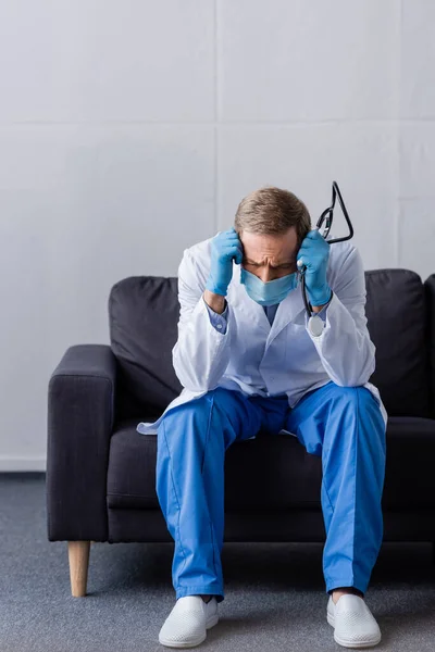 Втомлений зрілий лікар з мігрені тримає стетоскоп, сидячи на дивані — стокове фото