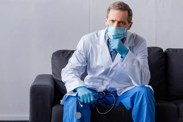 Mature médecin dans le masque médical tenant stéthoscope tout en étant assis sur canapé — Photo de stock