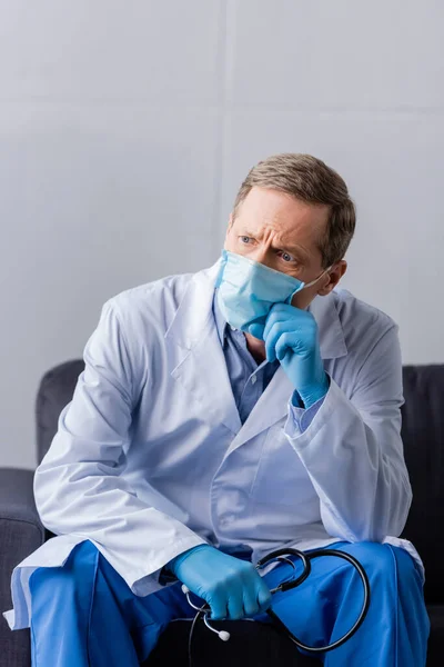 Задумчивый и зрелый врач в медицинской маске держит стетоскоп, сидя на диване — стоковое фото