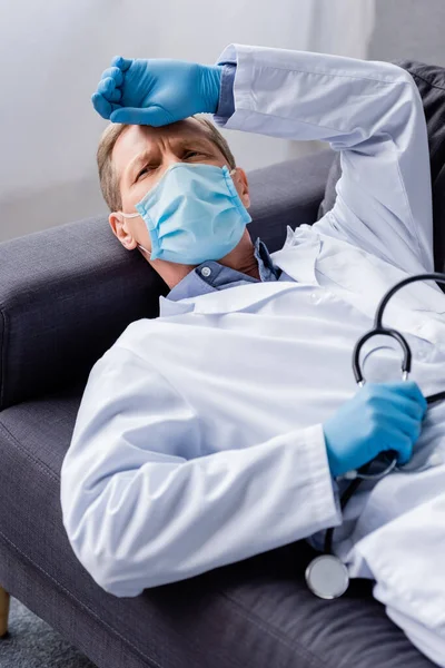 Tired and mature doctor in medical mask holding stethoscope while lying on sofa — Stock Photo