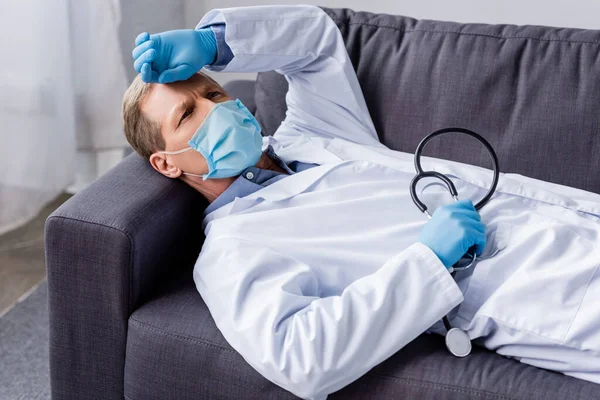 Измученный и зрелый врач в медицинской маске держит стетоскоп, лежа на диване — стоковое фото
