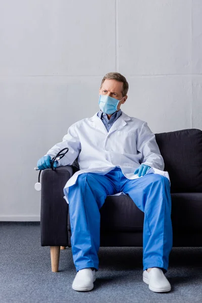 Зрелый врач в медицинской маске держит стетоскоп, сидя на диване — стоковое фото