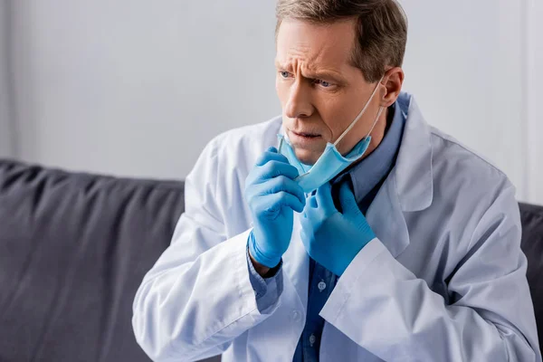 Upset mature doctor in latex gloves touching medical mask — Stock Photo