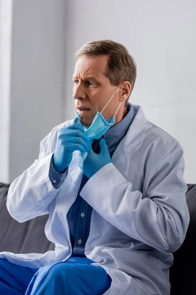 Médico triste y maduro en guantes de látex tocando máscara médica - foto de stock