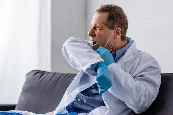 Médico maduro cansado en máscara médica y guantes de látex bostezando mientras está sentado en el sofá — Stock Photo
