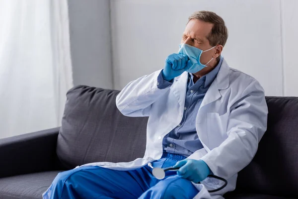 Tired and mature doctor in medical mask and latex gloves sitting on sofa — Stock Photo