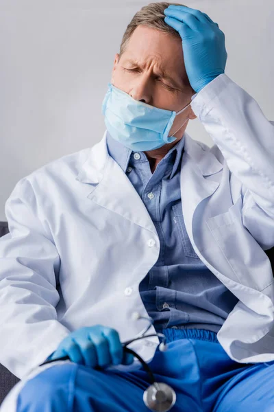 Selective focus of tired and mature doctor in medical mask and latex gloves touching head — Stock Photo