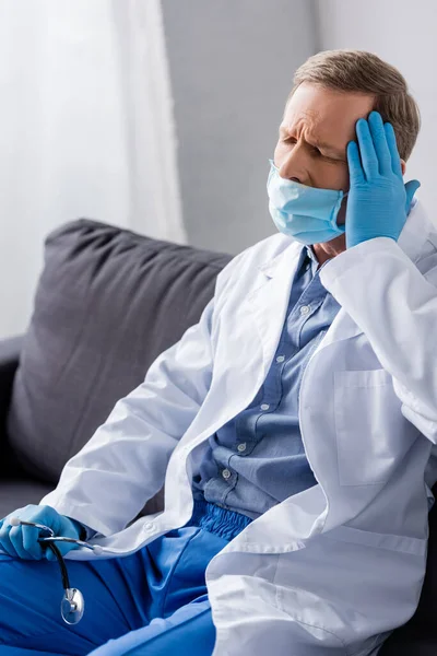 Tired and mature doctor with closed eyes touching head while sitting on sofa — Stock Photo