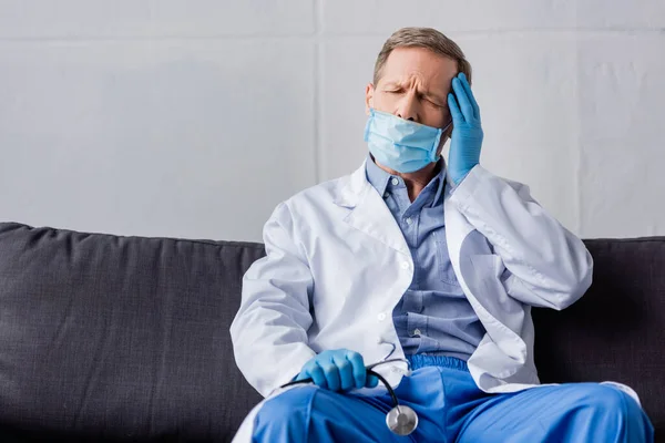 Tired mature doctor with closed eyes sitting on sofa and holding stethoscope — Stock Photo