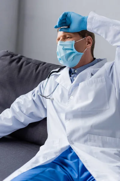 Tired and mature doctor in medical mask sitting on sofa — Stock Photo