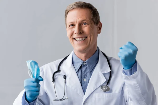 Happy and mature doctor in latex gloves holding medical mask on grey — Stock Photo