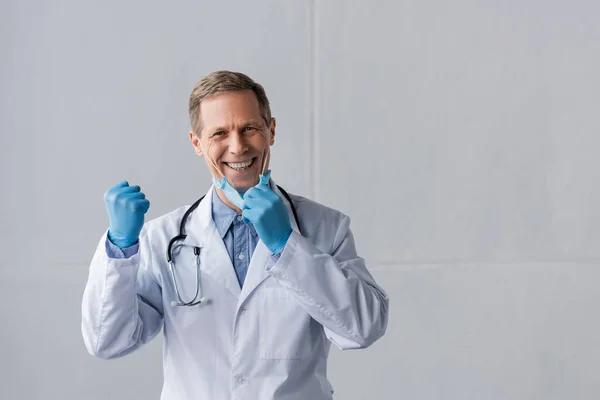 Médico feliz y maduro en guantes de látex y máscara médica con el puño cerrado en gris - foto de stock