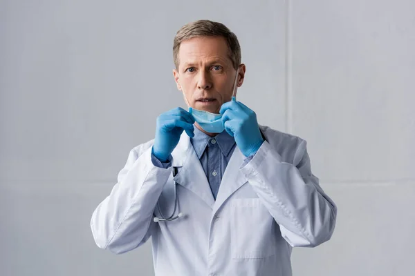 Shocked and mature doctor in latex gloves touching medical mask on grey — Stock Photo