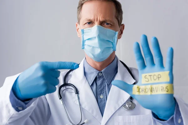 Enfoque selectivo del médico maduro en máscara médica apuntando con el dedo a la mano con letras de coronavirus stop aislado en gris - foto de stock
