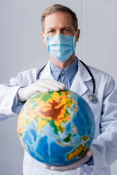 Вибірковий фокус зрілого лікаря в латексних рукавичках і медичній масці, що тримає глобус на сірому — стокове фото