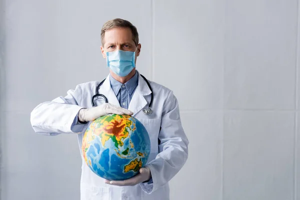 Зрілий лікар в латексних рукавичках і медичній масці тримає глобус на сірому — стокове фото