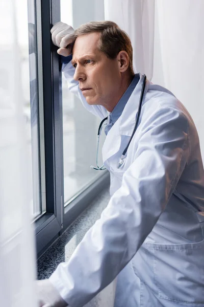 Tired middle aged doctor standing near window in clinic — Stock Photo