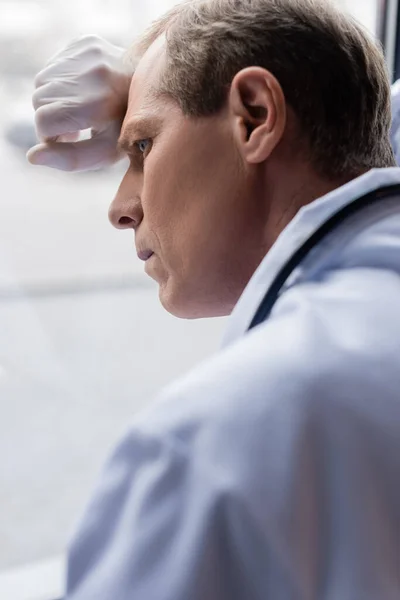 Selective focus of tired middle aged doctor standing near window in clinic — Stock Photo