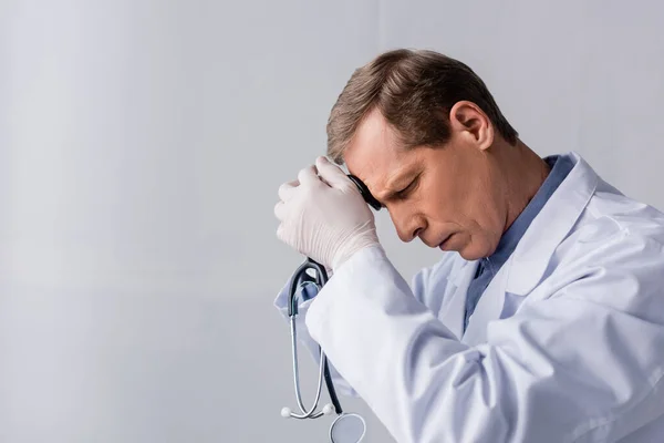 Tired and mature doctor with closed eyes holding stethoscope on grey — Stock Photo